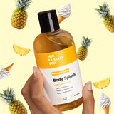 Hand holding Her Fantasy Box Pineapple Crème Body Splash bottle surrounded by pineapples and soft-serve ice cream cones on yellow background