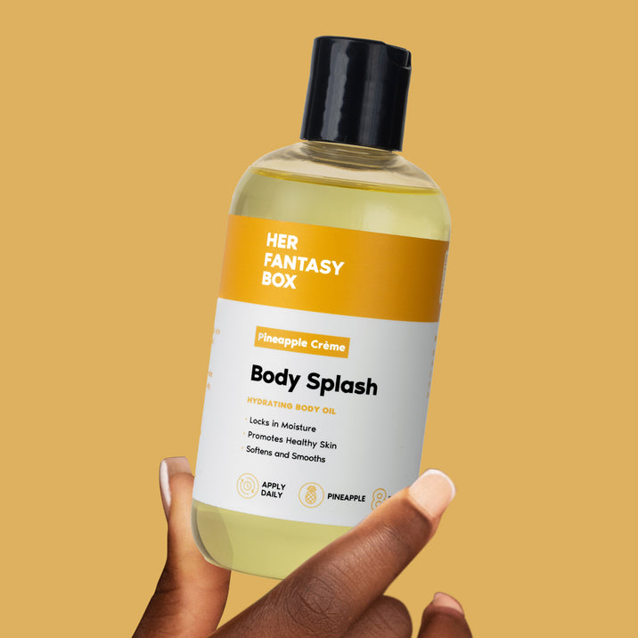 Hand holding Her Fantasy Box Pineapple Crème Body Splash hydrating body oil bottle against golden yellow background