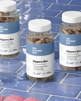 Three bottles of 'Slippery Box' capsules positioned on a blue tiled surface with water droplets around. 