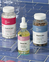 Her Fantasy Box bundle featuring Fresh & Clean suppositories, Renew Her oil, and Slippery Box displayed on blue wet tile