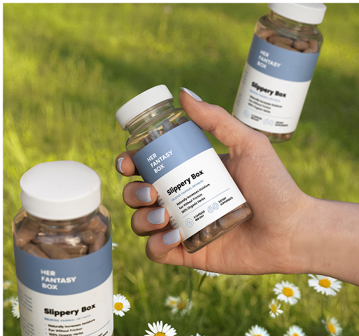 Hand holding a bottle of 'Her Fantasy Box Slippery Box' with grass and flowers in the background