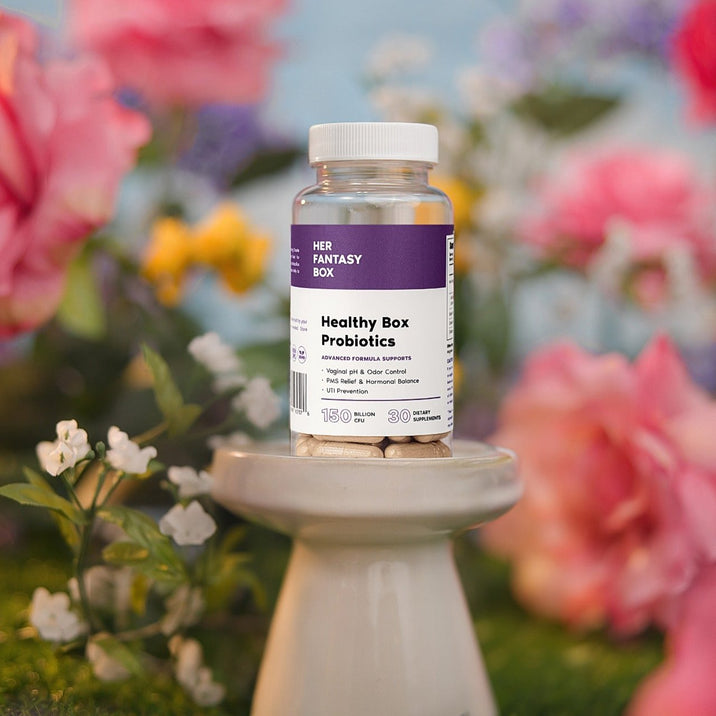 Her Fantasy Box Healthy Box Probiotics bottle displayed on a pedestal surrounded by pink and yellow flowers for vaginal health and UTI prevention